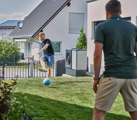 Heizung im Sommer modernisieren: Zei Männer spielen mit einem Ball im Garten, im Hintergrund ist ein Haus und eine Wärmepumpe zu sehen. 
