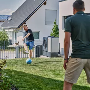 Heizung im Sommer modernisieren: Zei Männer spielen mit einem Ball im Garten, im Hintergrund ist ein Haus und eine Wärmepumpe zu sehen. 